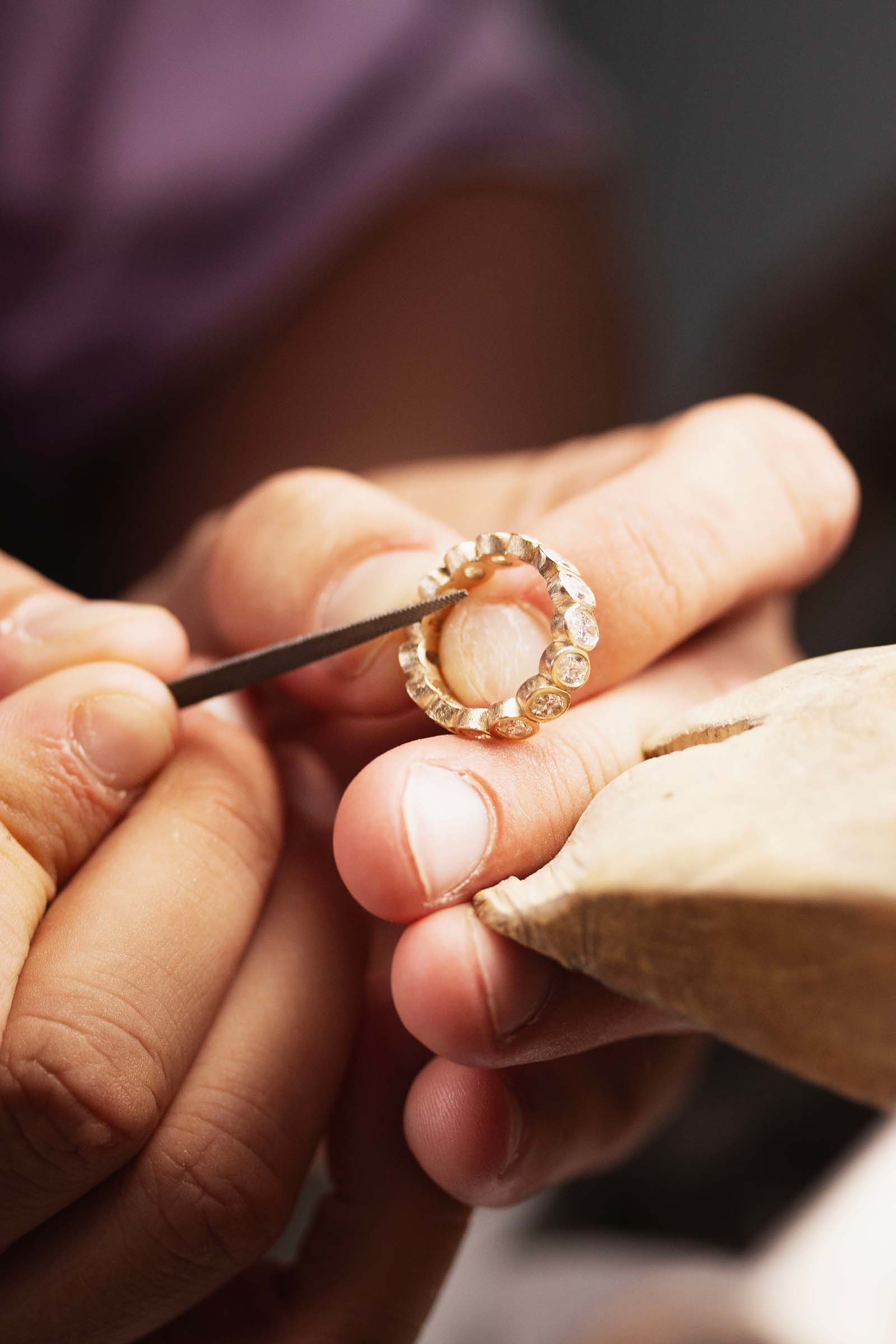 A close-up of a gold ring being held and worked on by a jeweler's hands, with a wooden loupe and a small metal tool visible against a softly blurred background.