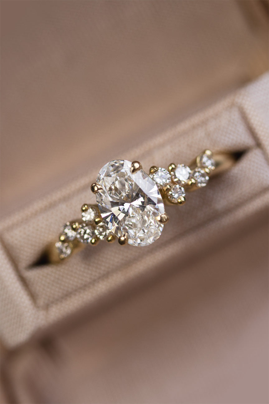 Close-up of a brilliant oval lab-grown diamond engagement ring with ten natural diamond accents on a gold band in a luxury box.