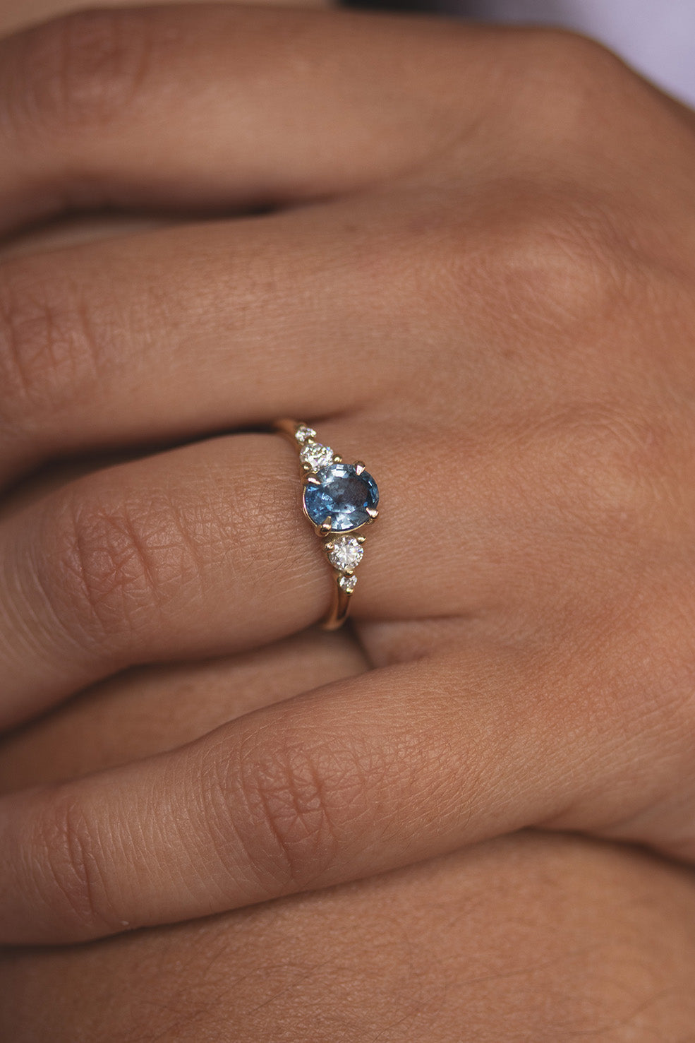 An oval blue sapphire engagement ring with diamond side stones worn on a finger, showing the vibrant ocean-blue color of the gem.